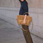 A woman stands on a city sidewalk against a stone wall, holding a large tan suede tote bag. She is wearing a navy long-sleeve top, olive green wide-leg pants, and bright red pointed shoes.