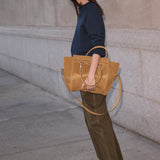 A woman stands on a city sidewalk against a stone wall, holding a large tan suede tote bag. She is wearing a navy long-sleeve top, olive green wide-leg pants, and bright red pointed shoes.