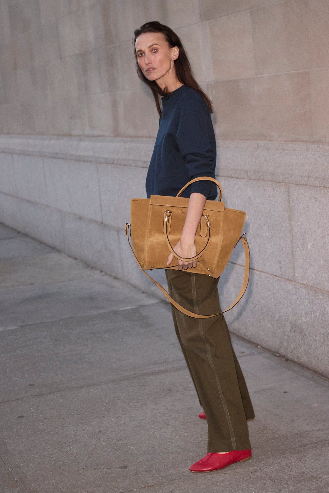 A woman stands on a city sidewalk against a stone wall, holding a large tan suede tote bag. She is wearing a navy long-sleeve top, olive green wide-leg pants, and bright red pointed shoes.