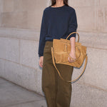 A woman stands against a light stone wall holding a large tan leather tote bag. She is wearing a navy long-sleeve top, olive green wide-leg pants, and bright red shoes.