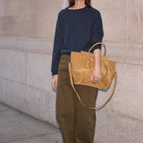 A woman stands against a light stone wall holding a large tan leather tote bag. She is wearing a navy long-sleeve top, olive green wide-leg pants, and bright red shoes.
