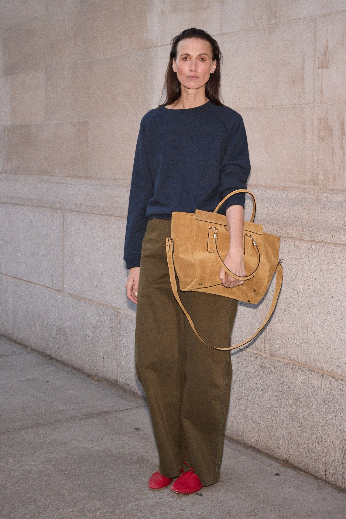 A woman stands against a light stone wall holding a large tan leather tote bag. She is wearing a navy long-sleeve top, olive green wide-leg pants, and bright red shoes.