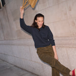 A woman in a navy long-sleeve top and olive green pants leans against a stone wall, holding a tan suede shoulder bag.