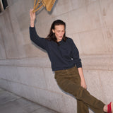 A woman in a navy long-sleeve top and olive green pants leans against a stone wall, holding a tan suede shoulder bag.