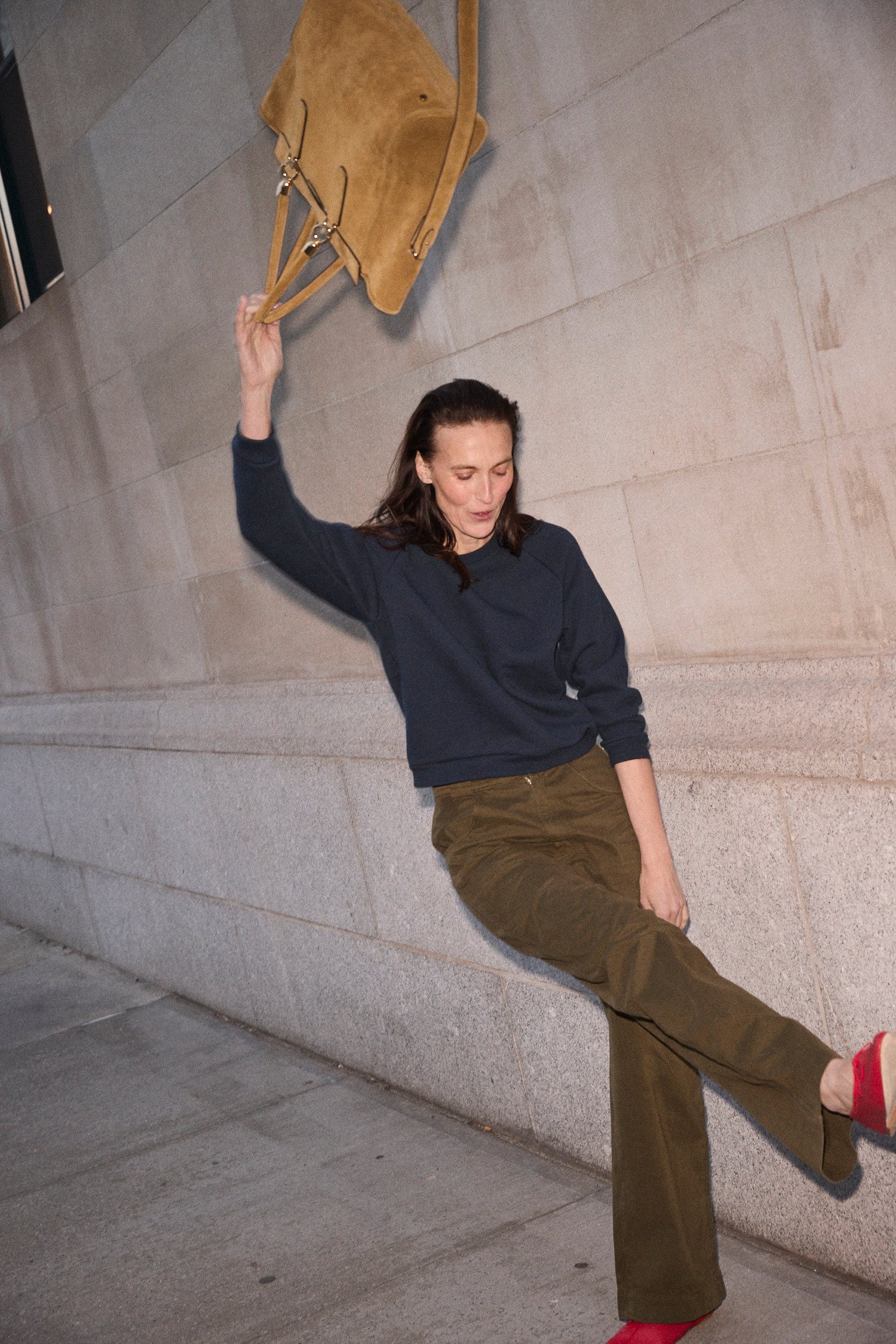A woman in a navy long-sleeve top and olive green pants leans against a stone wall, holding a tan suede shoulder bag.